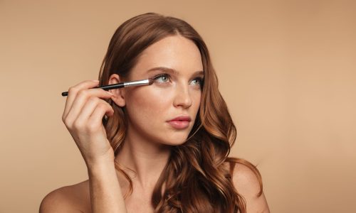 Beauty portrait of mystery ginger woman with long hair looking away while applying cosmetics with brush over cream background