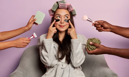 Pretty Korean woman points at eye patches, wears bathrobe, sits at home after shower, has healthy skin and complexion, isolated over purple wall. Beauty industry, cosmetology, skin care concept