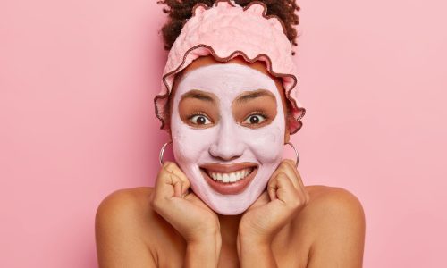 Isolated shot of young girl stands with mud rejuvenation mask, holds hands under chin, looks gladfully at camera, stands with bare shoulders, has well cared complexion, regular beauty treatments